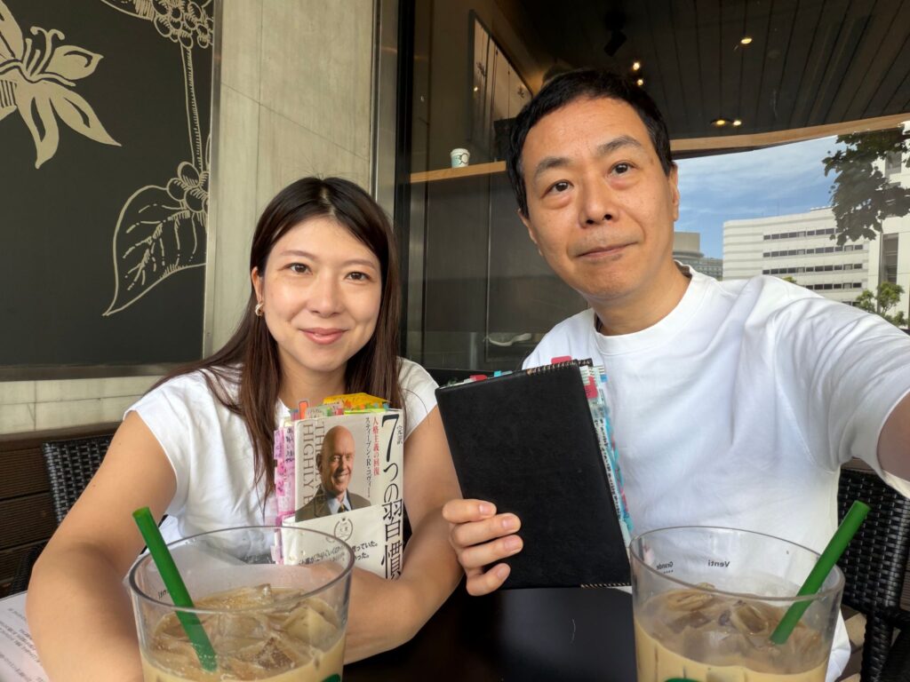 付箋がたくさん貼られた『完訳 7つの習慣』と手帳を手に、カフェのテラス席で実践会を行う二人