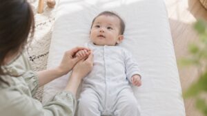 白い布団に仰向けの赤ちゃんの小さな手を、母の両手がそっと包んでいる
