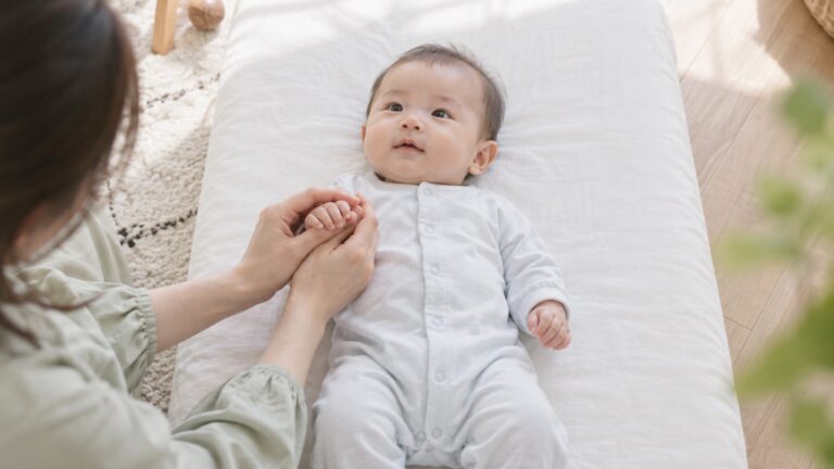 白い布団に仰向けの赤ちゃんの小さな手を、母の両手がそっと包んでいる