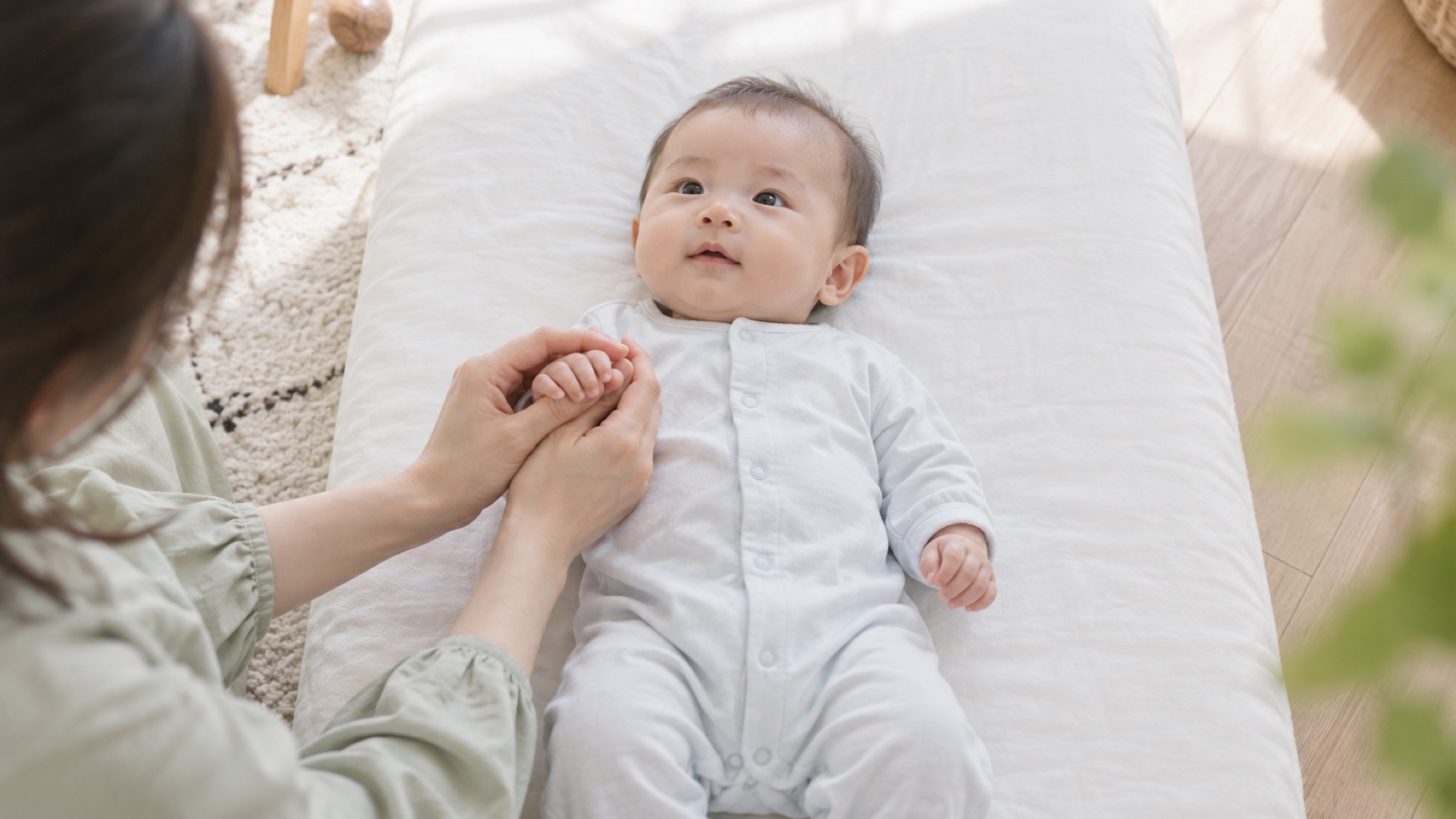 白い布団に仰向けの赤ちゃんの小さな手を、母の両手がそっと包んでいる